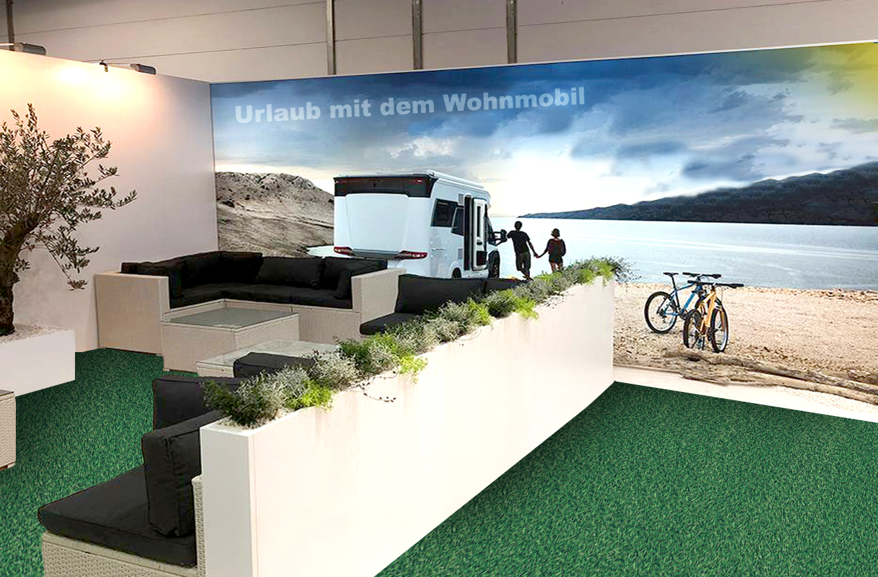 Grüner Kunstrasen als Bodenbelag auf einem Messestand mit Lounge-Möbeln, Pflanzkübeln und großer Wohnmobil-Urlaubswand im Hintergrund
