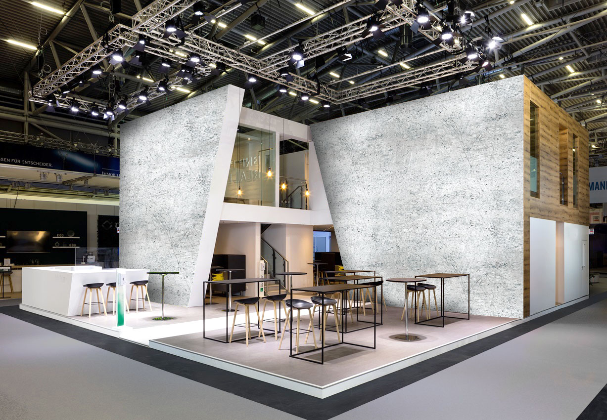 Moderner zweistöckiger Messestand mit großen Wänden in Betonoptik, Holzverkleidung, Beleuchtung, Bar und Lounge-Möbeln in einer Messehalle