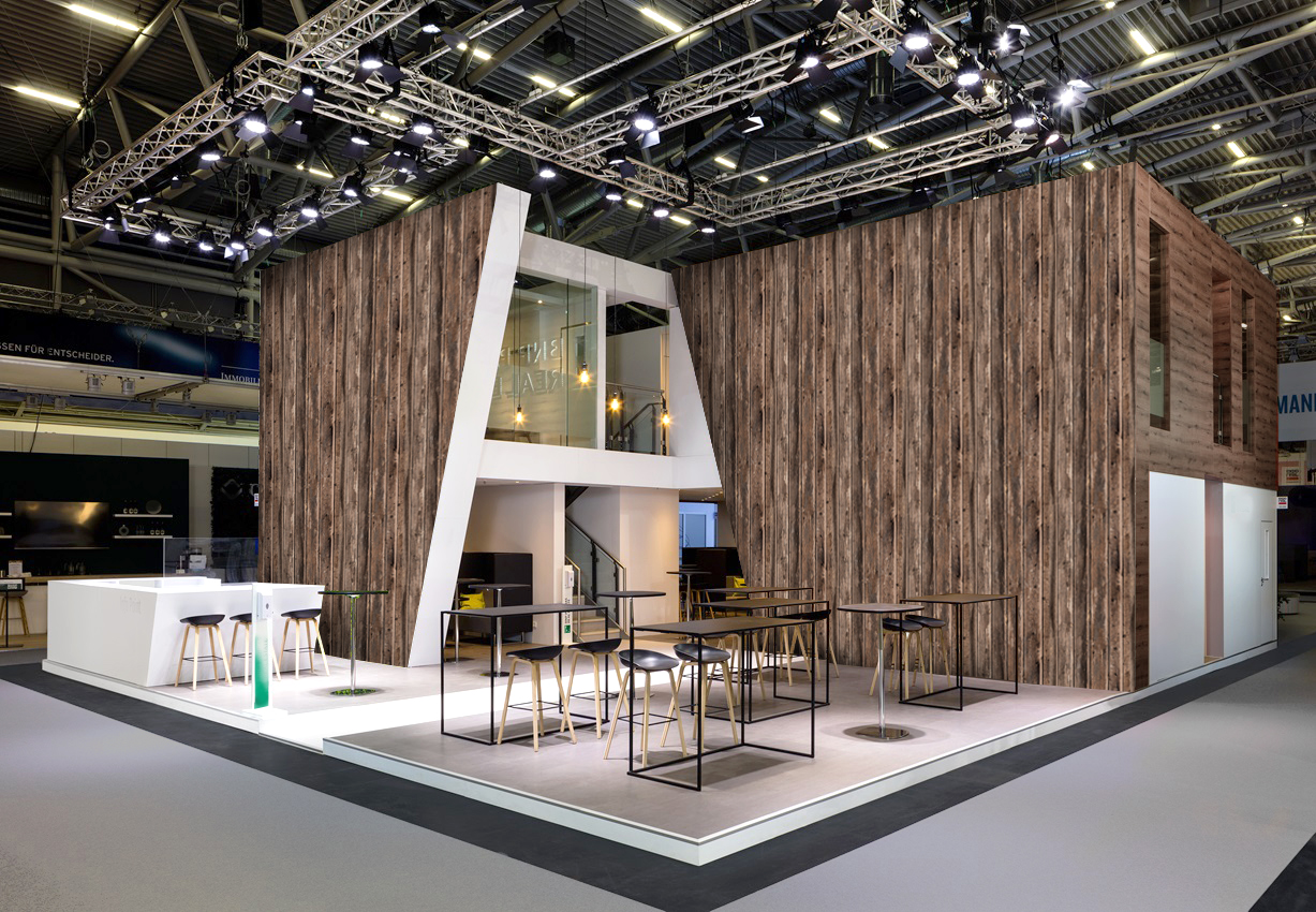 Großer moderner Messestand mit rustikaler Holzverkleidung, heller Theke, Tischen, Barhockern und professioneller Deckenbeleuchtung in einer Messehalle