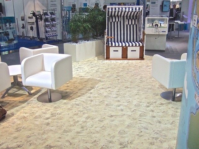 Messestand mit sandfarbener Bodenfläche im Strandlook, weißen Loungesesseln, Pflanzkübel und Strandkorb als einladende Präsentationsfläche