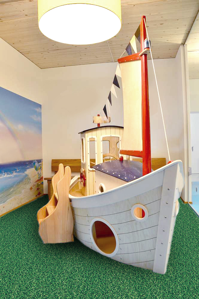 Großes Holz-Spielschiff für den Innenbereich mit Segel, Mast, Bullaugen und Rutsche auf grünem Spielteppich im Kinderzimmer.