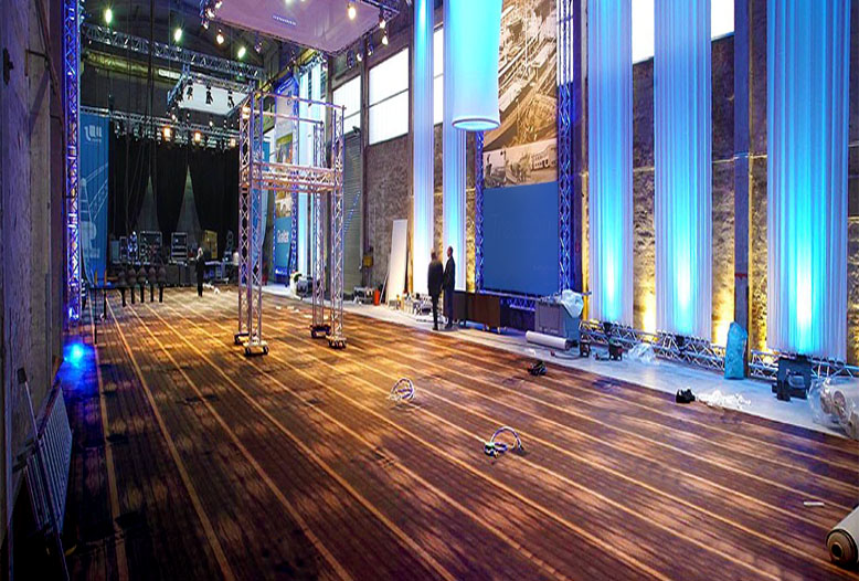 Große Eventhalle mit Teppichboden, Traversen, Lichttechnik und blau beleuchteten Säulen beim Aufbau einer professionellen Veranstaltung.